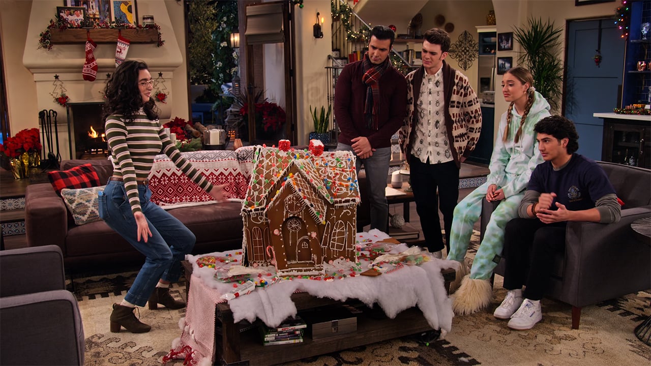 A still from Ashley Garcia: Genius in Love: Christmas showing a group of teens in a living room looking at a big gingerbread house on the coffee table