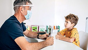 Photo of a kid during a speech therapy session. The speech therapist is wearing a face mask and face shield while holding up flashcards with big letters printed on them