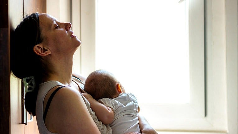 A stressed mother sits on the floor with her baby sleeping on her chest and her head against a cupboard with her eyes closed
