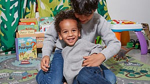 Brothers in grey thermal long sleeve shirts and jeans snuggle in their play room-one has curly hair and one has straight hair for a story about them not being able to get their hair cut at the same salon
