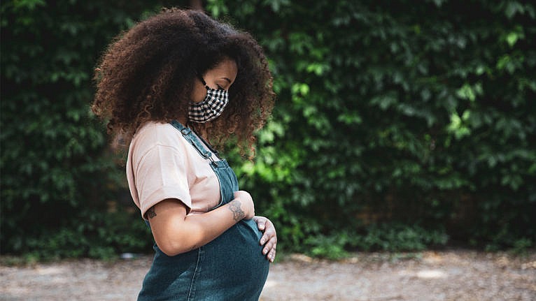 Side profile of a pregnant woman with an afro in a black and white gingham mask holding her belly in a pink tshirt and overalls in front of a wall of greenery