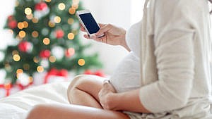 a pregnant woman sits cross legged on her bed holding her belly and a phone in the other hand with a christmas tree in the background