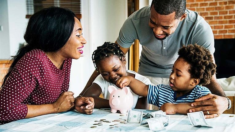 A photo of a family sitting at the dining table, putting money into a piggy bank together