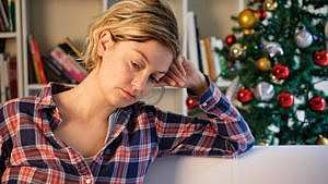 Photo of a woman looking sad while sitting on a couch in front of a Christmas tree