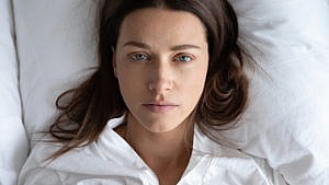 A photo of a woman lying in bed that only shows her from the neck up. She has a sad expression on her face