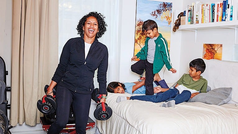Photo of a mom carrying dumbbells while her kids play on the bed nearby