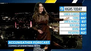 A weather forecaster holds up her son against a forecast screen with a chart of weather forecasts in california to her right