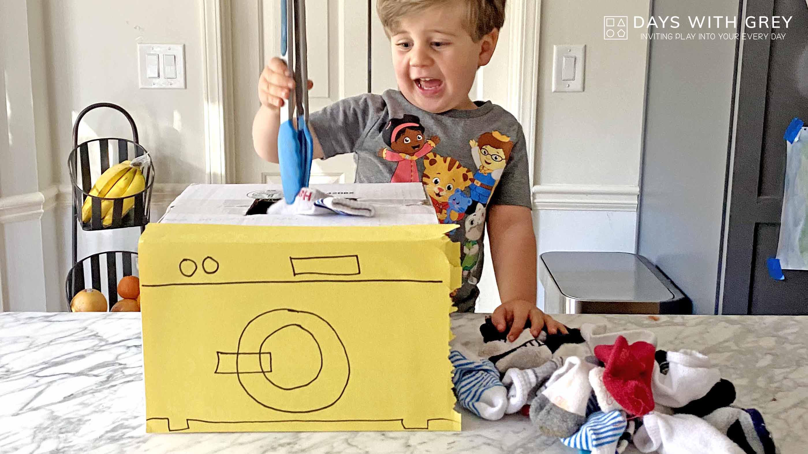 A young boy puts socks into a cardboard washing machine using tongs for a story on toddler activities you can set up the night before