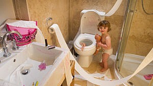 photo of a toddler in a diaper making a big mess in the bathroom. Toilet paper is unraveled, There are make-up drawings all over the sink and toilet and a doll is sticking out of the toilet bowl