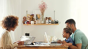 Photo of a couple working at the kitchen table. One of them carries a baby sucking on a pacifier while they work on a laptop