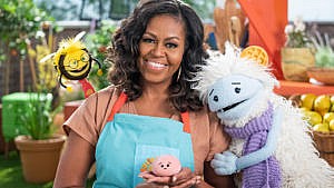 Promo photo from Michelle Obama's new netflix show Waffles and Mochi showing Michelle posing with three puppets. One is a yeti wearing a scarf with a waffle on their head, another is a bee wearing glasses and a necktie and the last is a little pink ball of mochi