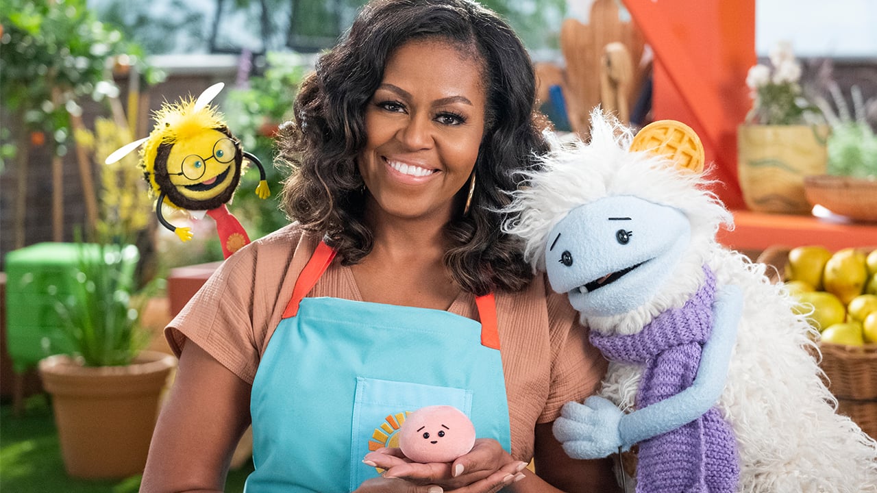 Promo photo from Michelle Obama's new netflix show Waffles and Mochi showing Michelle posing with three puppets. One is a yeti wearing a scarf with a waffle on their head, another is a bee wearing glasses and a necktie and the last is a little pink ball of mochi
