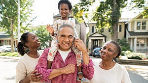 photo of a elderly couple standing outside on the street with their child and grandchild. the grandchild is sitting on grandpa's shoulders