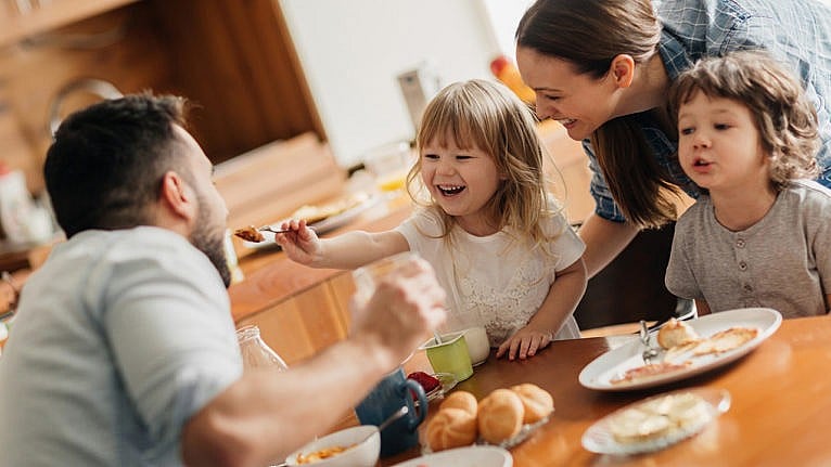 Photo of a family laughing at the dining table while a kid feeds the dad