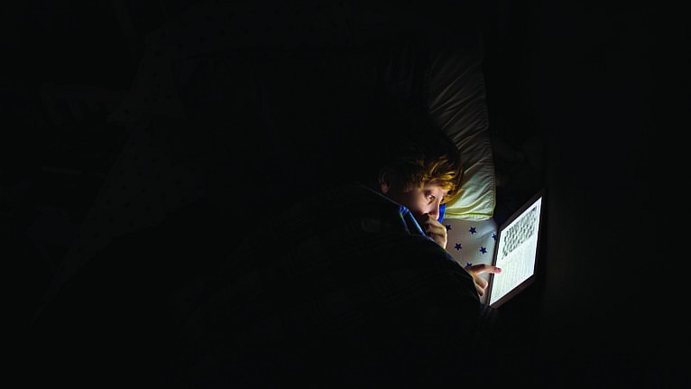 a kid lies in bed in a dark room with the glow of a tablet illuminating his face for a story on night owls