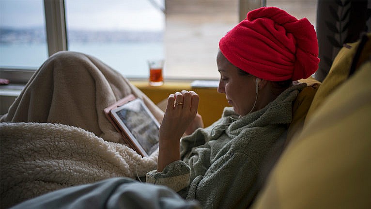 Photo of a woman lounging in a robe with her hair up in a towel looking at a tablet