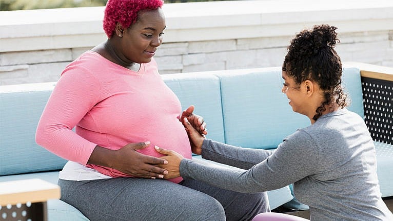 Photo of a pregnant woman sitting on an outdoor sofa being checked by her doula who is kneeling on the floor holding the woman's baby bump