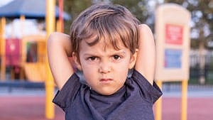 a little boy at the playground with his arms over his head staring at the camera with an angry look