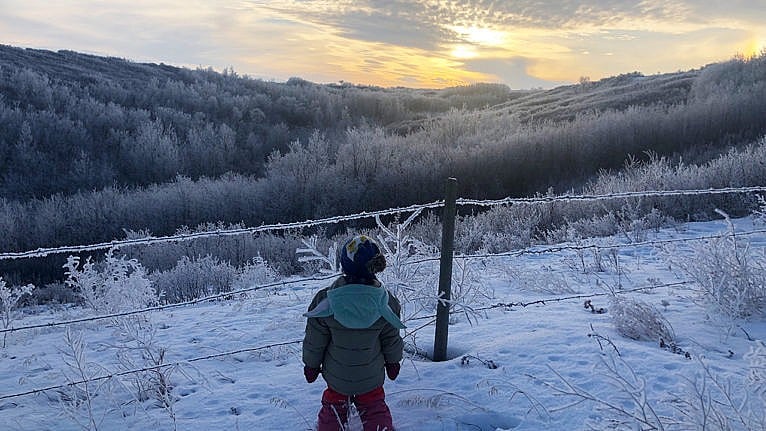 photo of a kid standing by a fence that is covered in ice looking out at a sun peeking thourgh the clouds over the hills