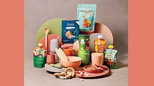 An array of packaged baby foods by Gerber, Baby Gourmet, Heinz and more with pretty bowls, plates and utensils in peach, clay, lime green and rose colours
