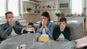 a young family who are all sick sit on the couch blowing their noses