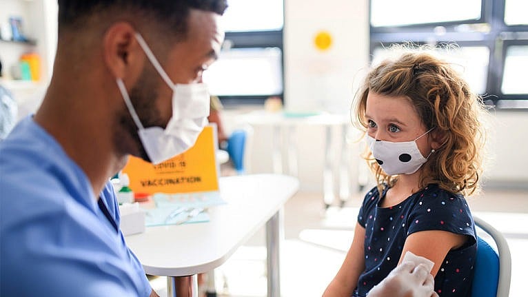Photo of young kid wearing a mask as a masked healthcare worker preps their arm for a vaccine shot