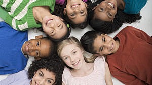 A diverse group of elementary age students are lying in a circle together and are smiling while looking up at the camera.