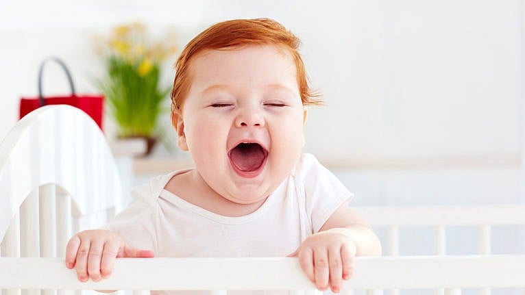 A baby stands in their crib with their mouth open looking gleeful