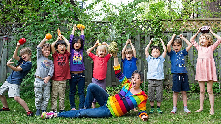 A group of kids holding produce with an adult in a rainbow sweater in front of them.