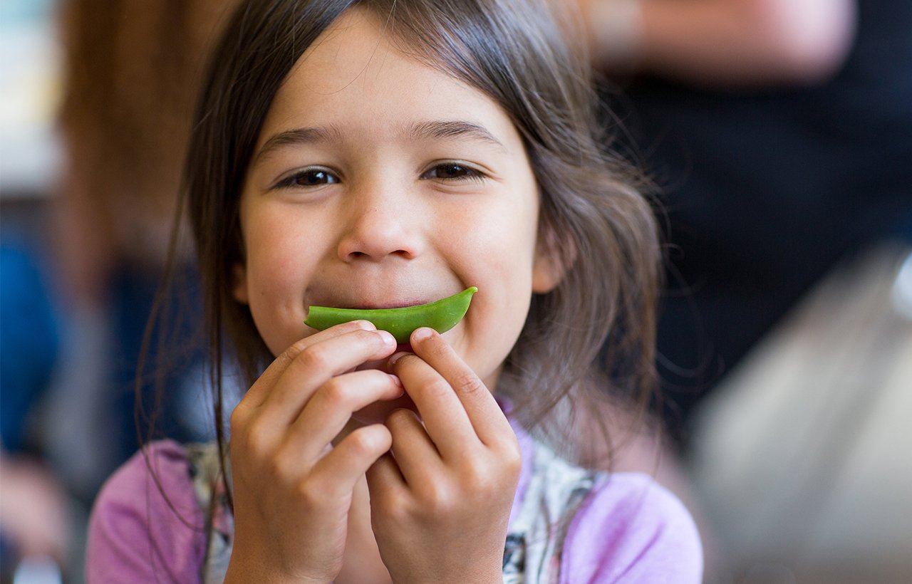 A child holds a pea pod to their mouth while smiling.