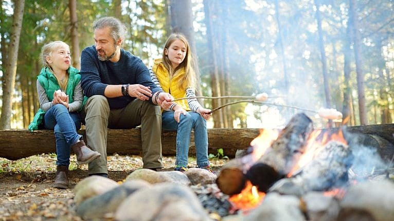 two young girls roasting marshmallows with their dad in a forest