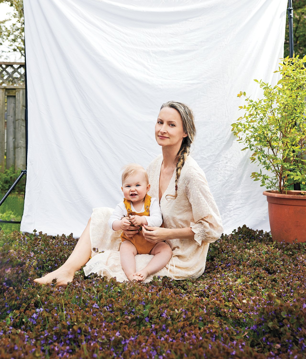 a mother sits outside with her baby on her lap and a white sheet hanging behind them