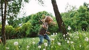 photo of a young kid running through a grassy patch of dandelions holding a stick
