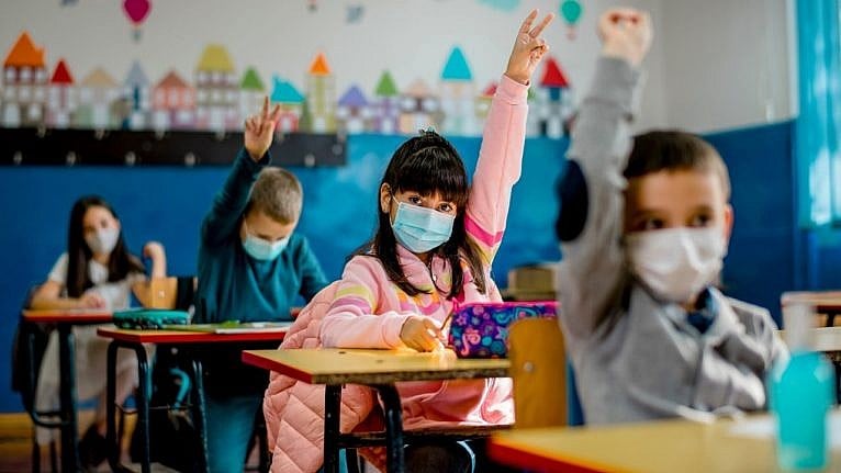 kids sitting at their school desks with their hands up wearing masks
