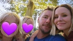 kristen bell and dax shepard pose with their two daughters outside under a tree with lights