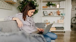 A young woman sits on her couch and browses the web on her laptop at home