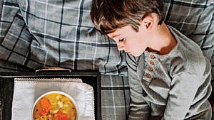 a young boy lies in bed looking sick with a bowl of chicken soup on a tray beside him