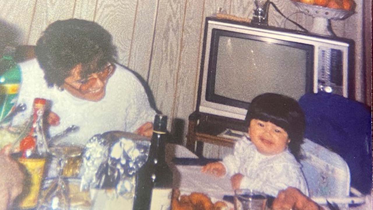 Photo of Melissa sitting in a baby seat on the right and her mother smiling at her on the left. They are sitting at a table with a television in the background.