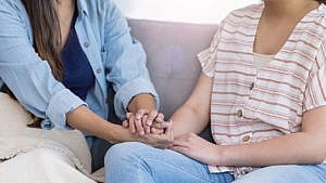 A photo of a parent in a blue button up shirt holding their kids hand. The kid is in a short sleeve stripped shirt. They are both sitting on a couch.