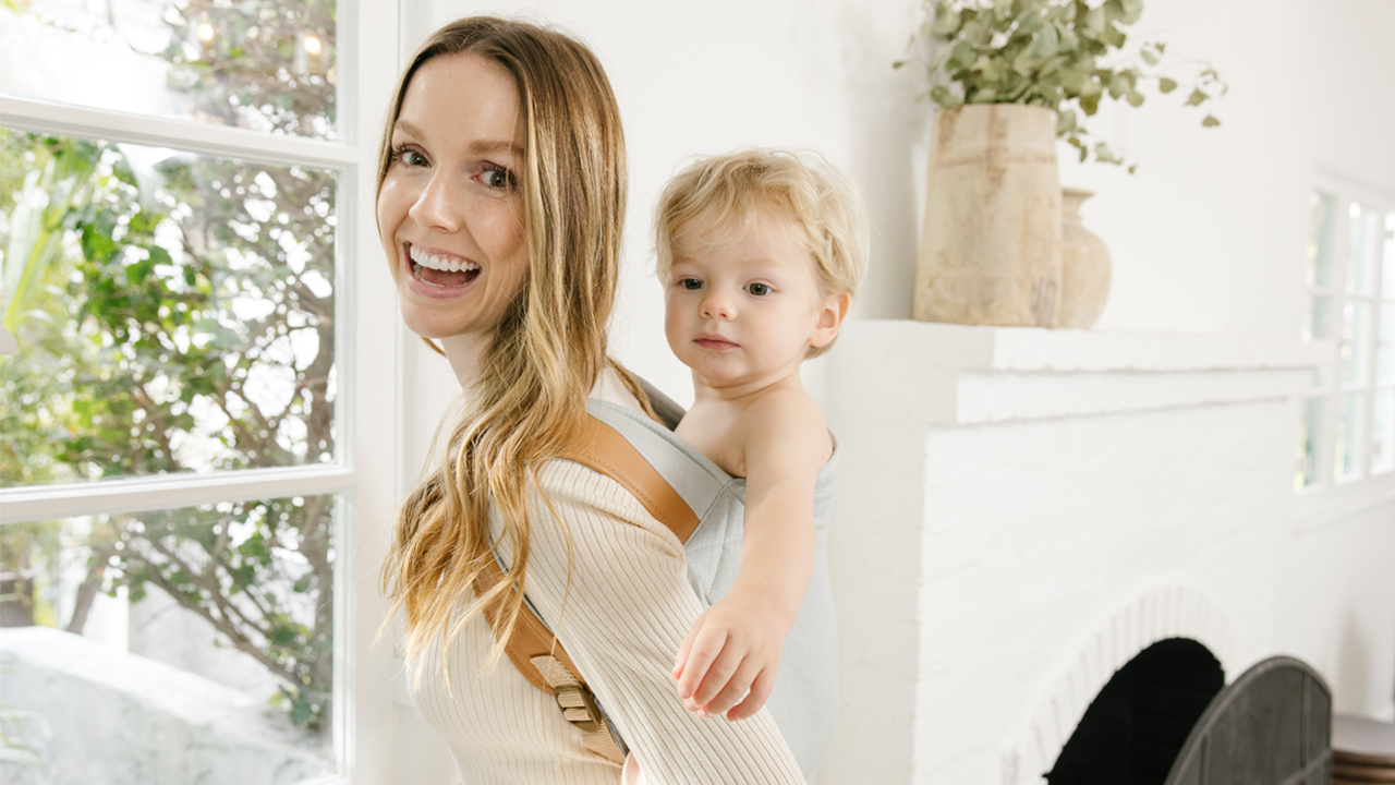 A photo of a parent in a white shift and blond hair, wearing a baby carrier on their back. Their baby is inside the carrier. They are in a white room with a window.