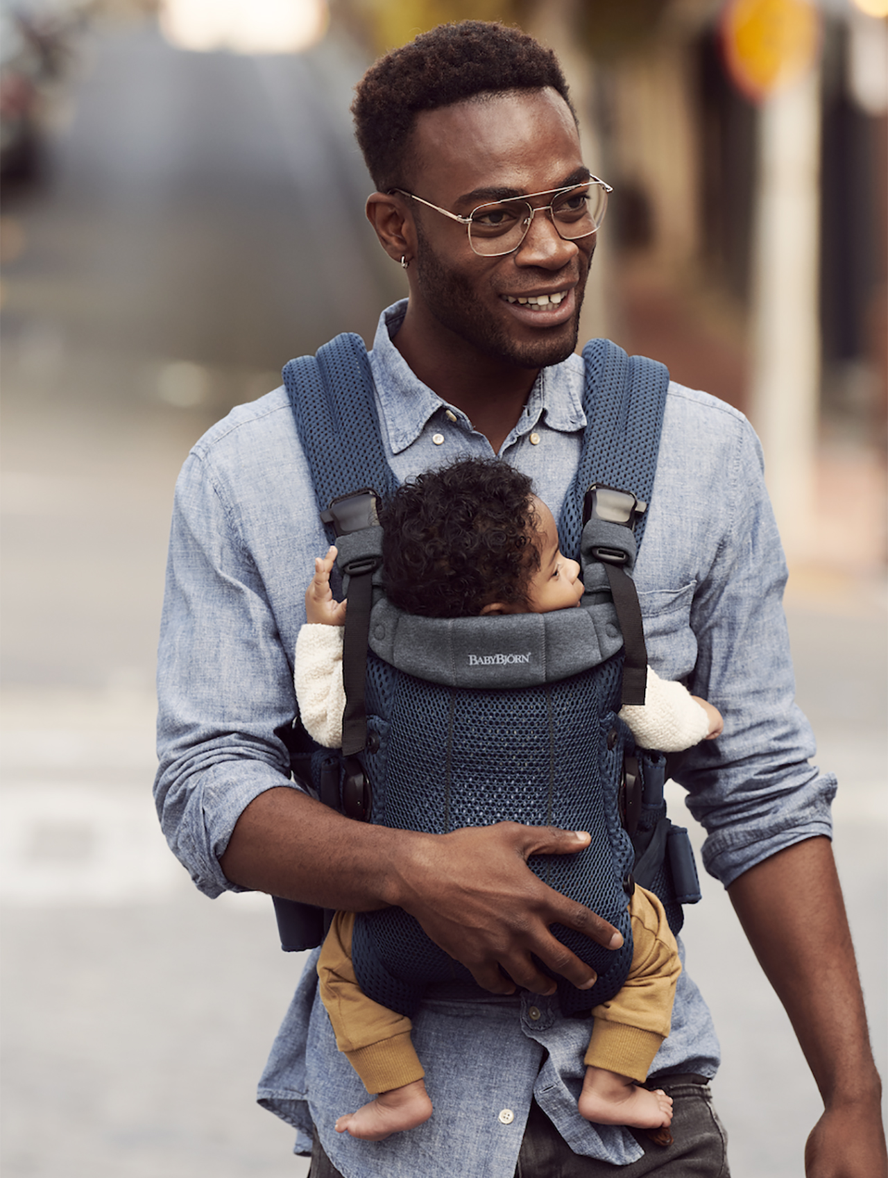 Een foto van een ouder in een blauw overhemd met knoopsluiting en een blauwe BabyBjörn Harmony-draagzak met hun kind erin,