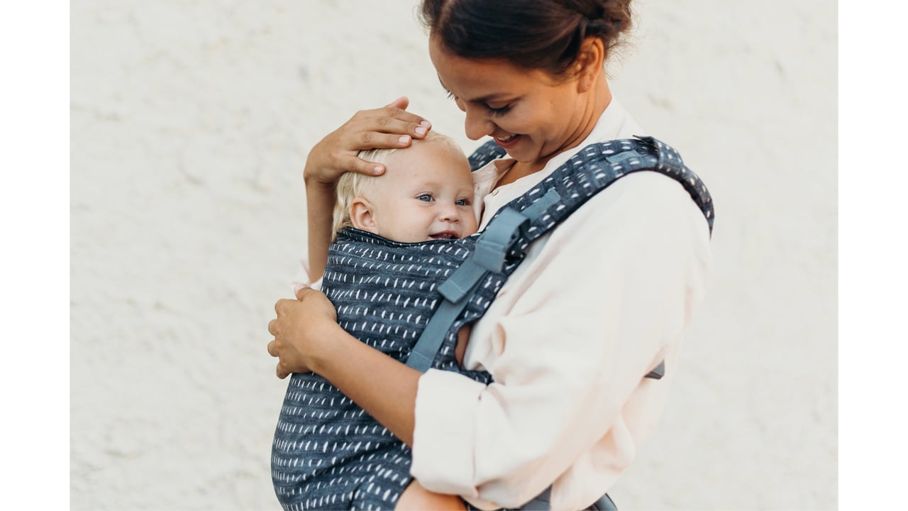 A photo of a parent in a white shirt wearing a blue Boba X baby carrier with white spots. They are carrying their smiling baby inside.