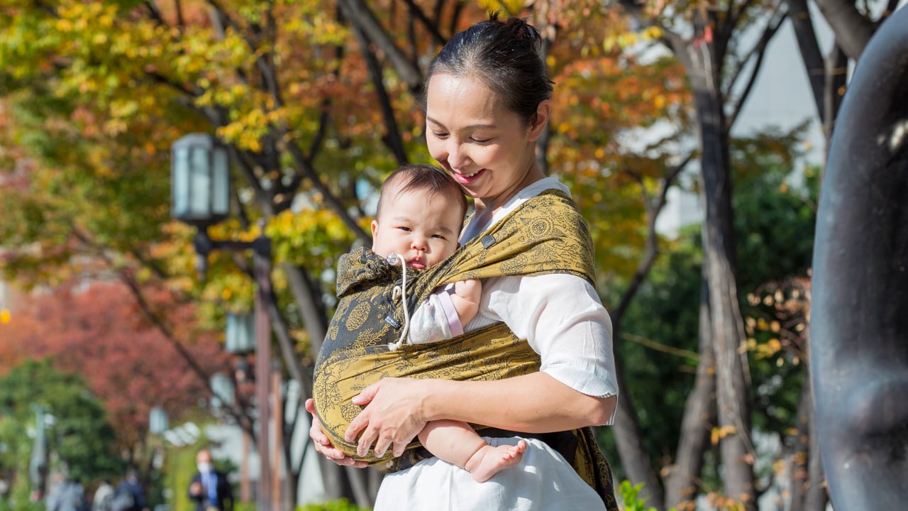 A photo of a parent in a white shirt wearing a green and grey patterned Didyklick with their baby inside. They are outside with yellow and orange leafed trees in the background.