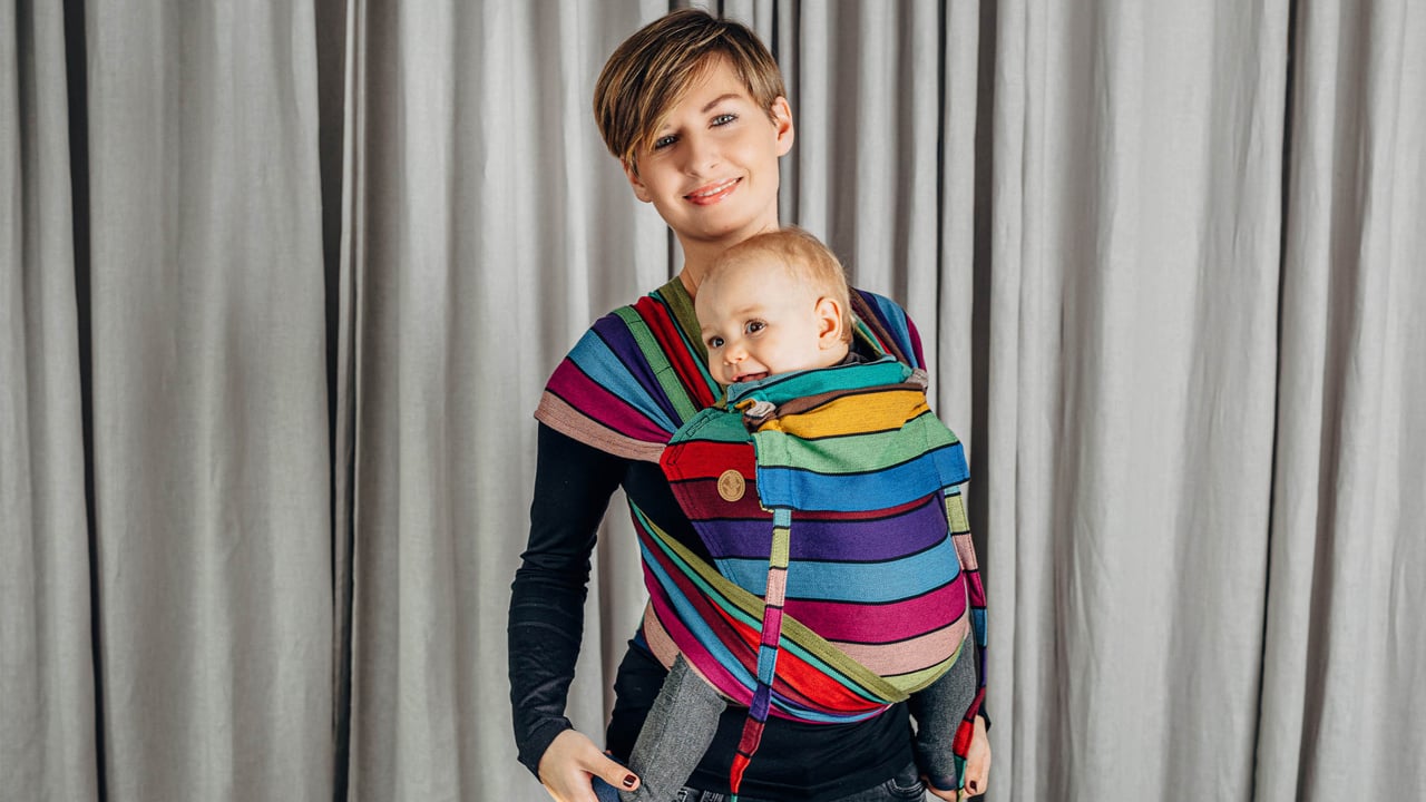 A parent wearing a multi-coloured WrapTai baby carrier with their baby inside. They are standing against a light grey curtain.