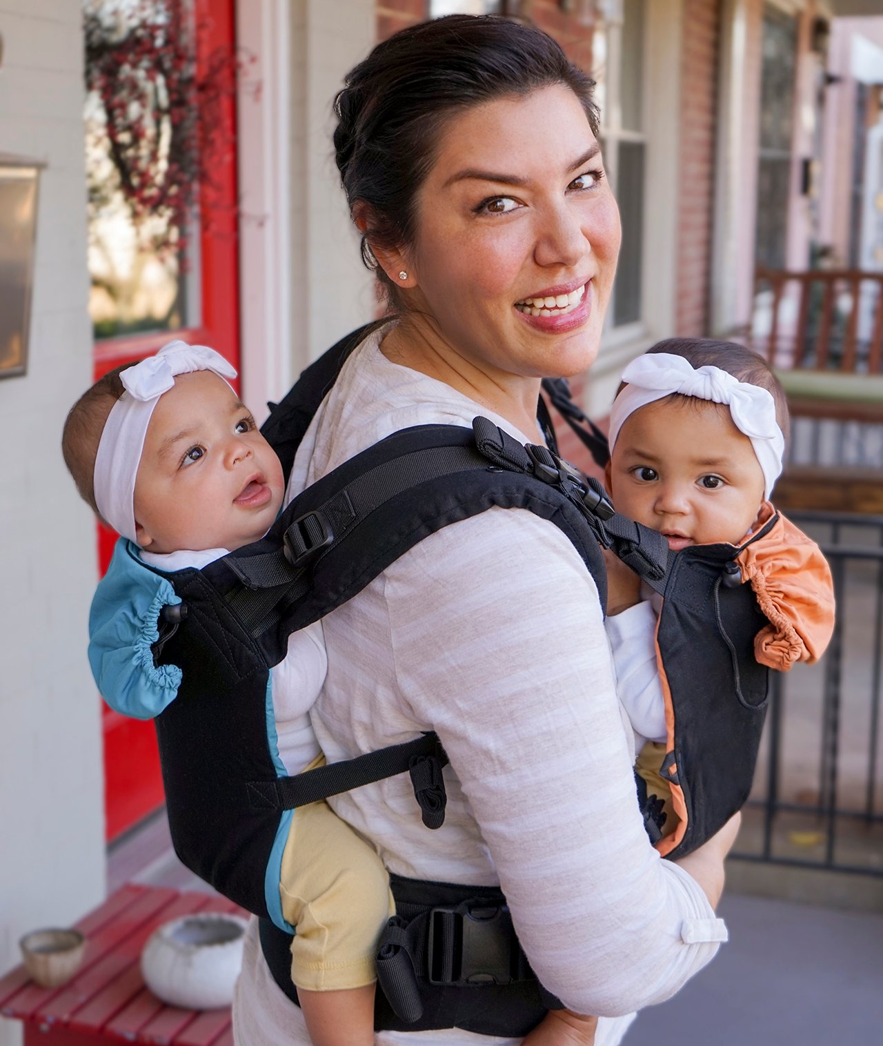 A photo of a parent in a white and pink striped top wearing a twin baby carrier. They have one baby on their chest and another on their back. Both babies are wearing bow headbands.