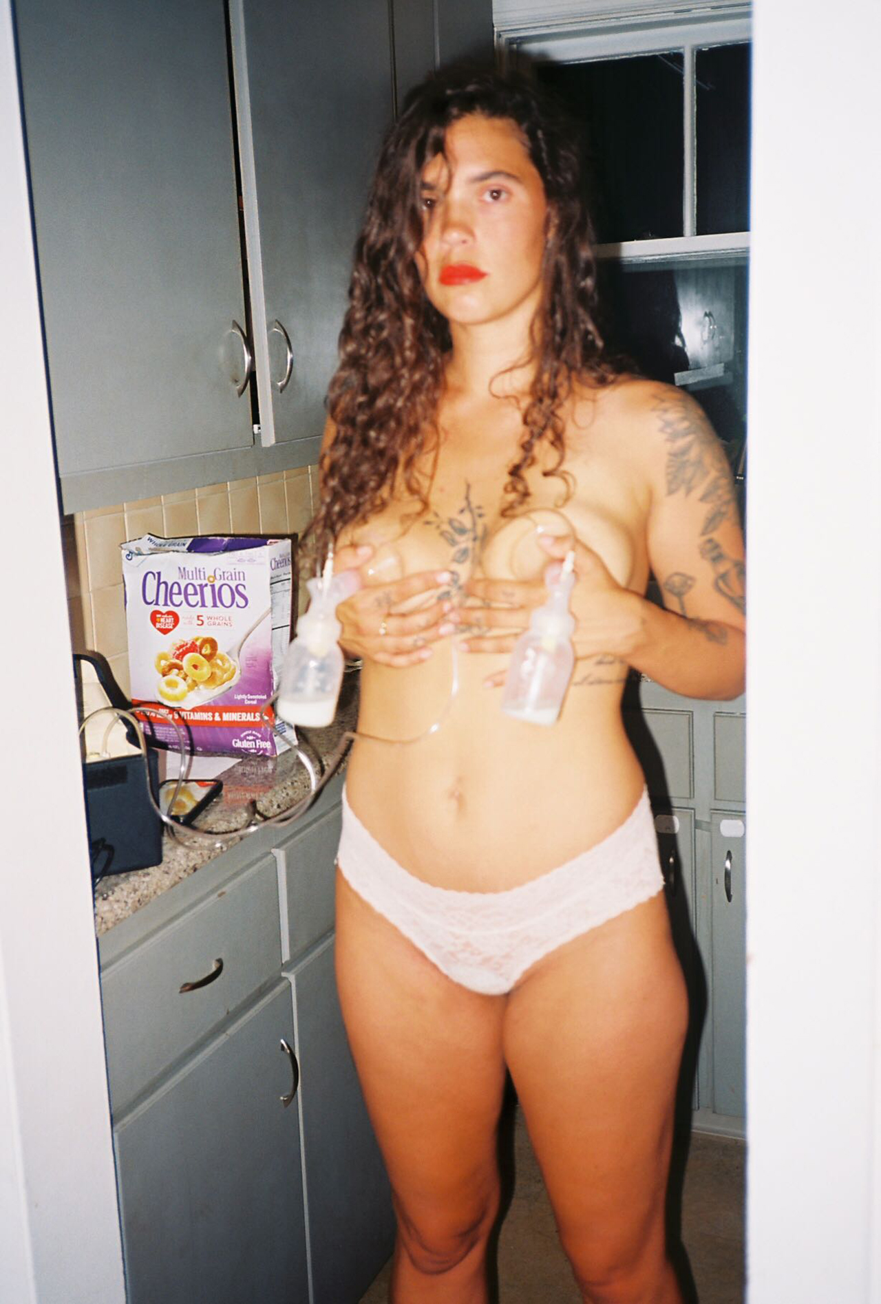 a woman stands in her kitchen pumping breastmilk with a box of cheerios in the background from the life after birth coffee table book about the postpartum experience