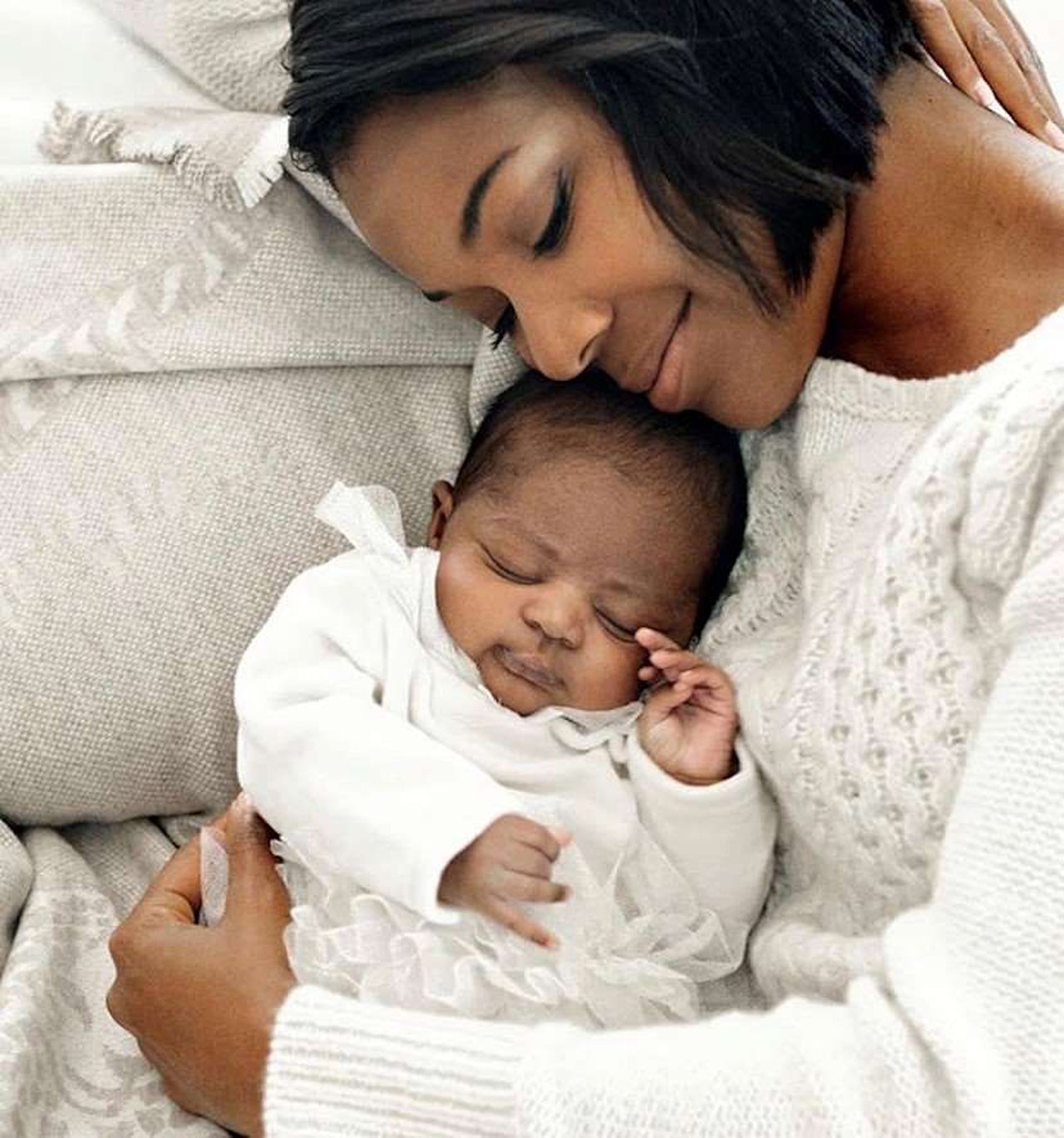 a woman in a white sweater smiles as she holds her newborn baby to her chest