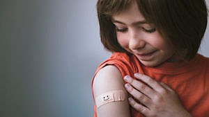 Photo of a young kid with a bandaid on their shoulder after getting a shot. the bandaid has a happy face drawn on it in black marker