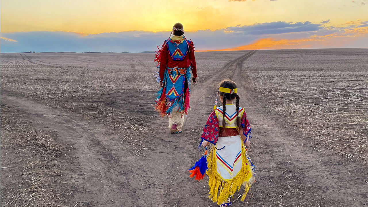 An adult and a child dressed in ceremonial Indigenous dresses walk towards the sunset on the beach