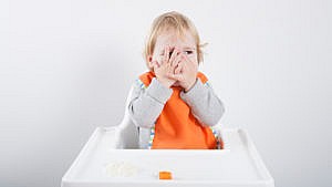 a toddler sits in a high chair with their hands over their face blocking food